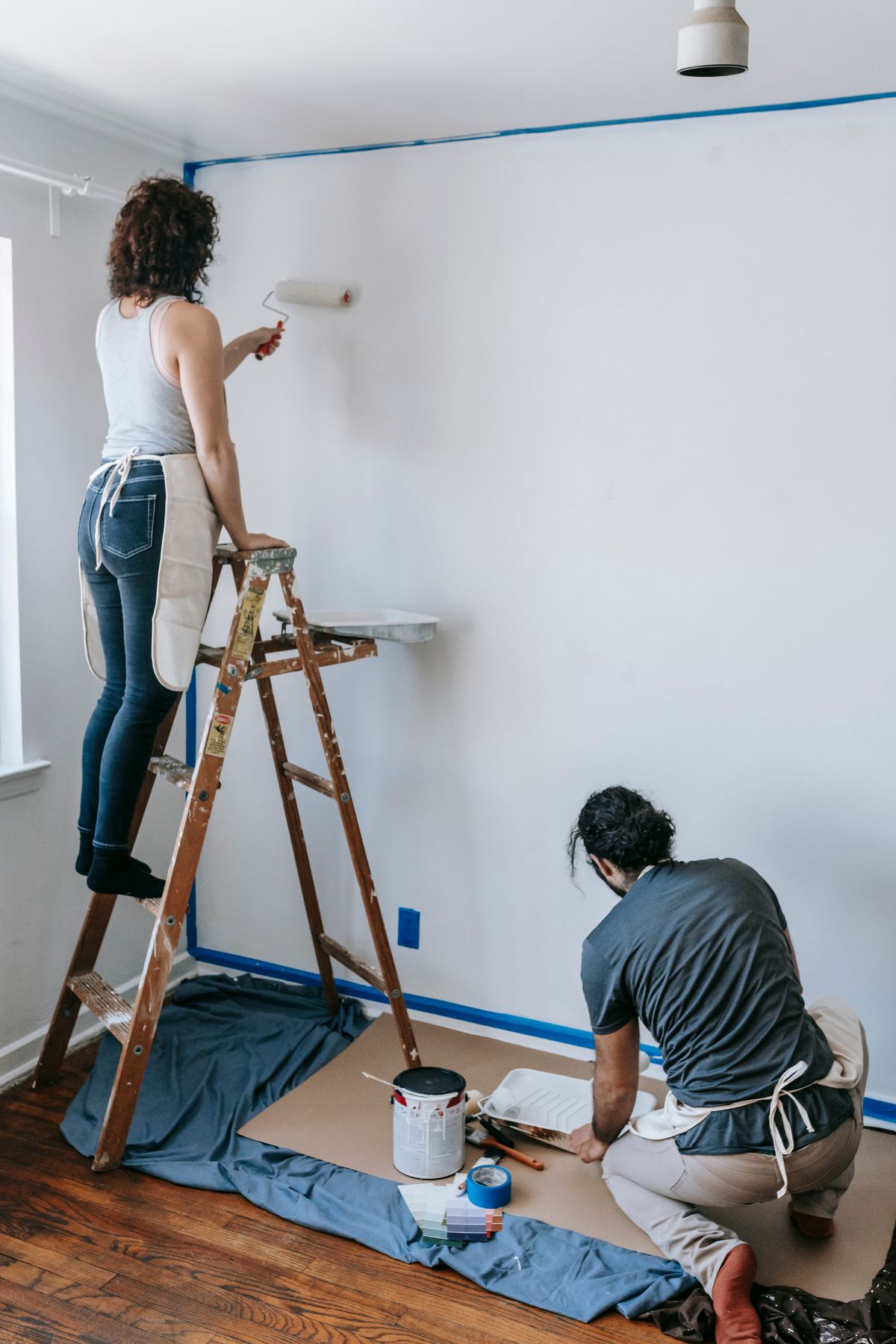 Two decorators painting a bright living room, one cutting in from a ladder